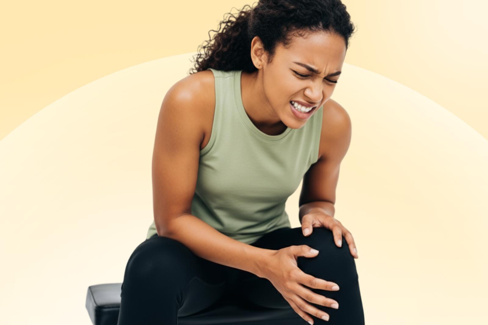 Woman in a green tank top sitting, grimacing in pain, holding her knee, against a soft yellow background.