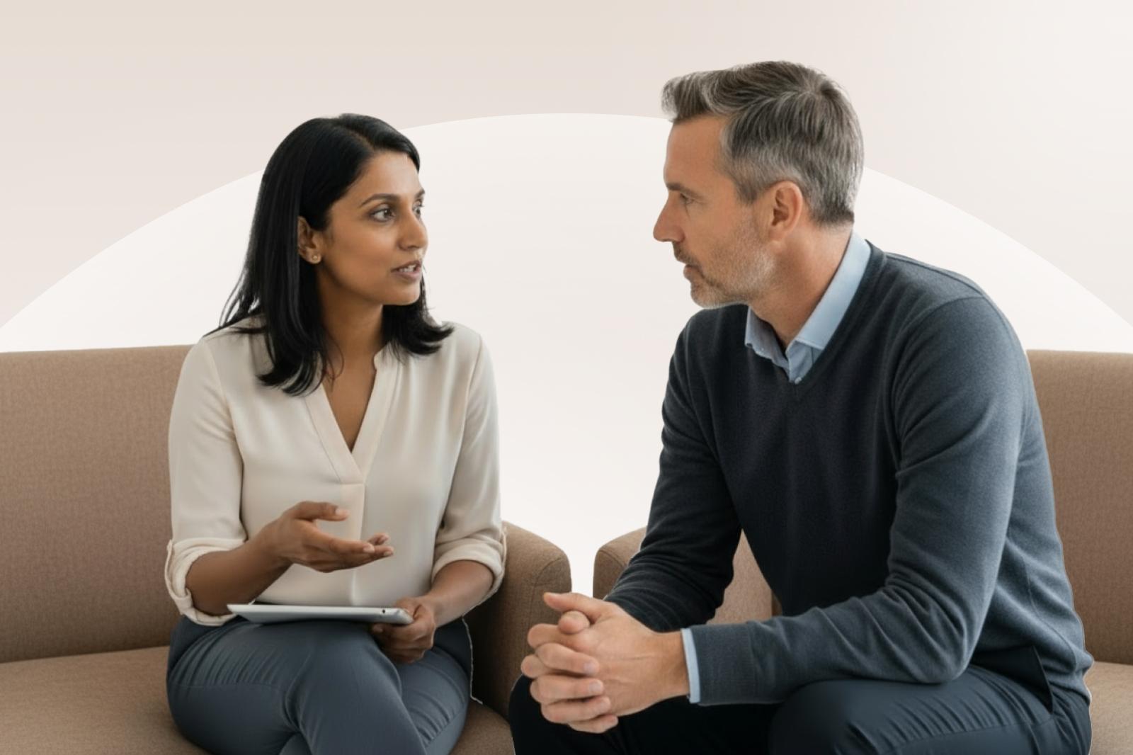 A woman and a man sit on a couch, engaged in a conversation. She holds a tablet, and both appear focused and attentive.