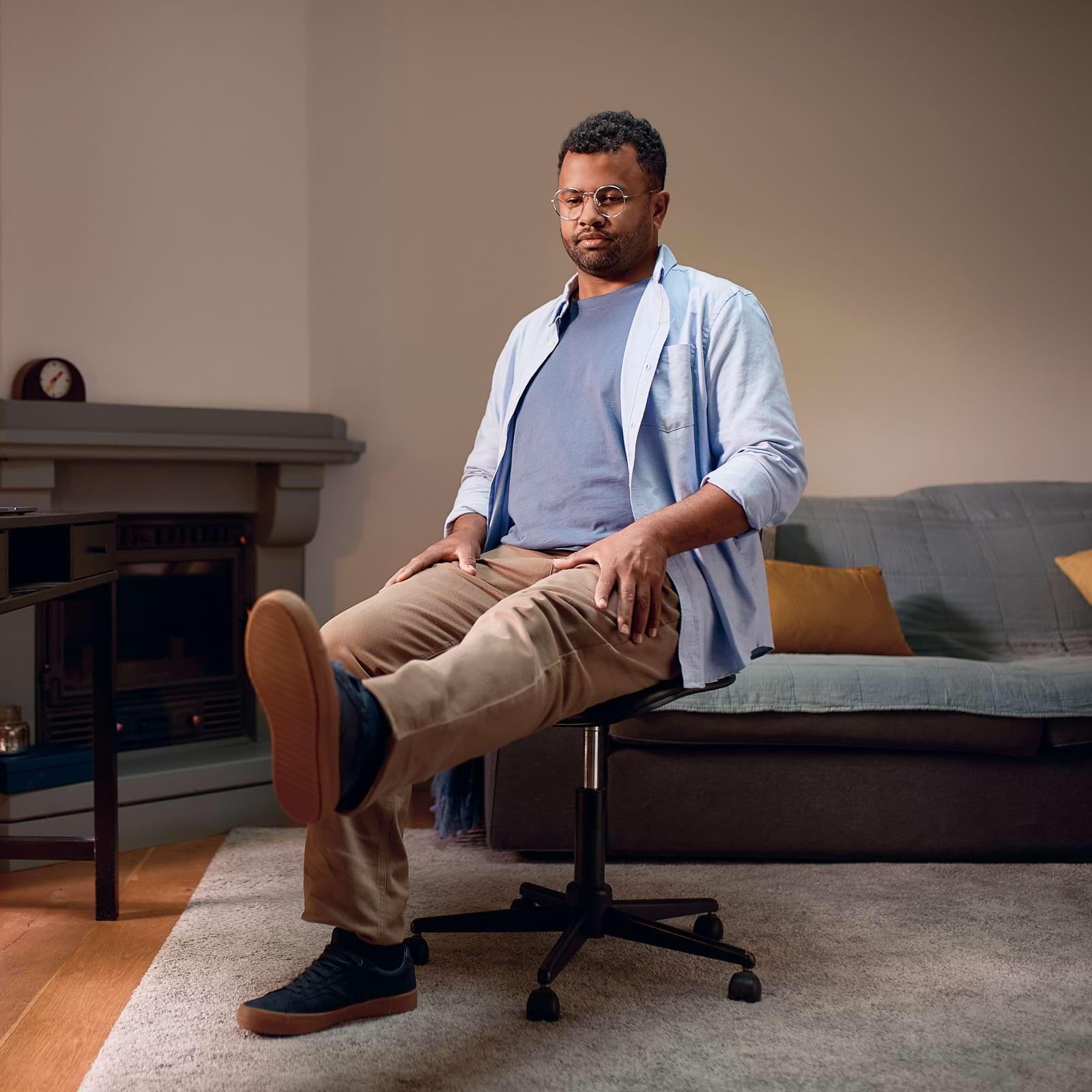 Man sitting on an office chair in a living room, one leg raised, wearing a blue shirt and beige pants, with a sofa and fireplace in the background.