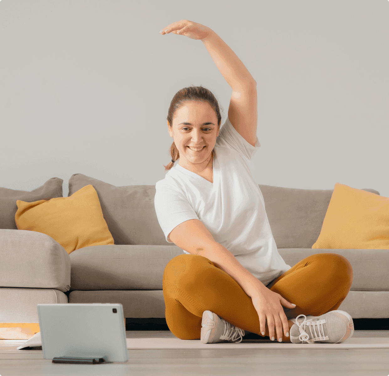 Woman sitting cross-legged on a floor, stretching with one arm raised, in front of a tablet. Couch with yellow pillows in the background.