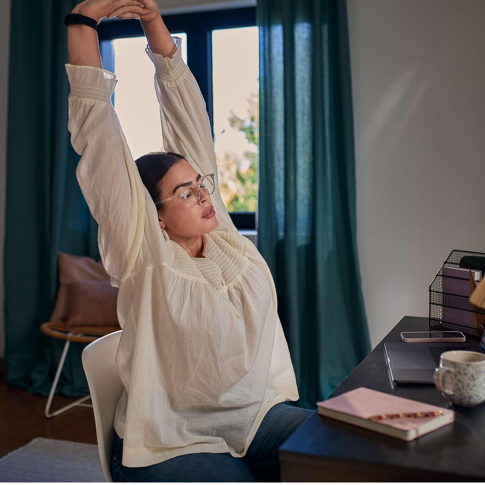 Woman stretching at a desk in a cozy room, wearing glasses and a white blouse. A laptop, notebook, and mug are on the desk.