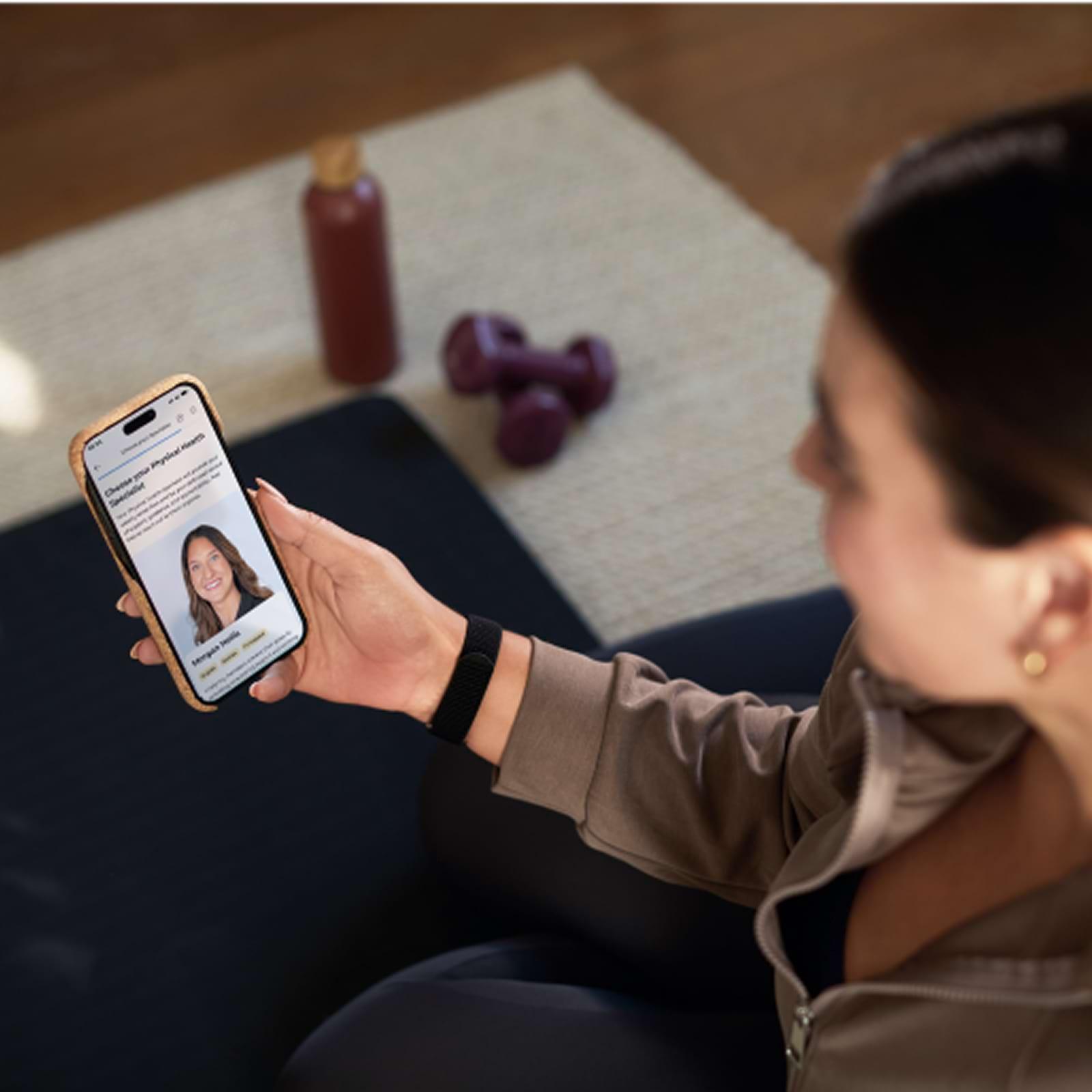 Person holding a smartphone displaying a profile, sitting on a yoga mat with dumbbells and a water bottle nearby.