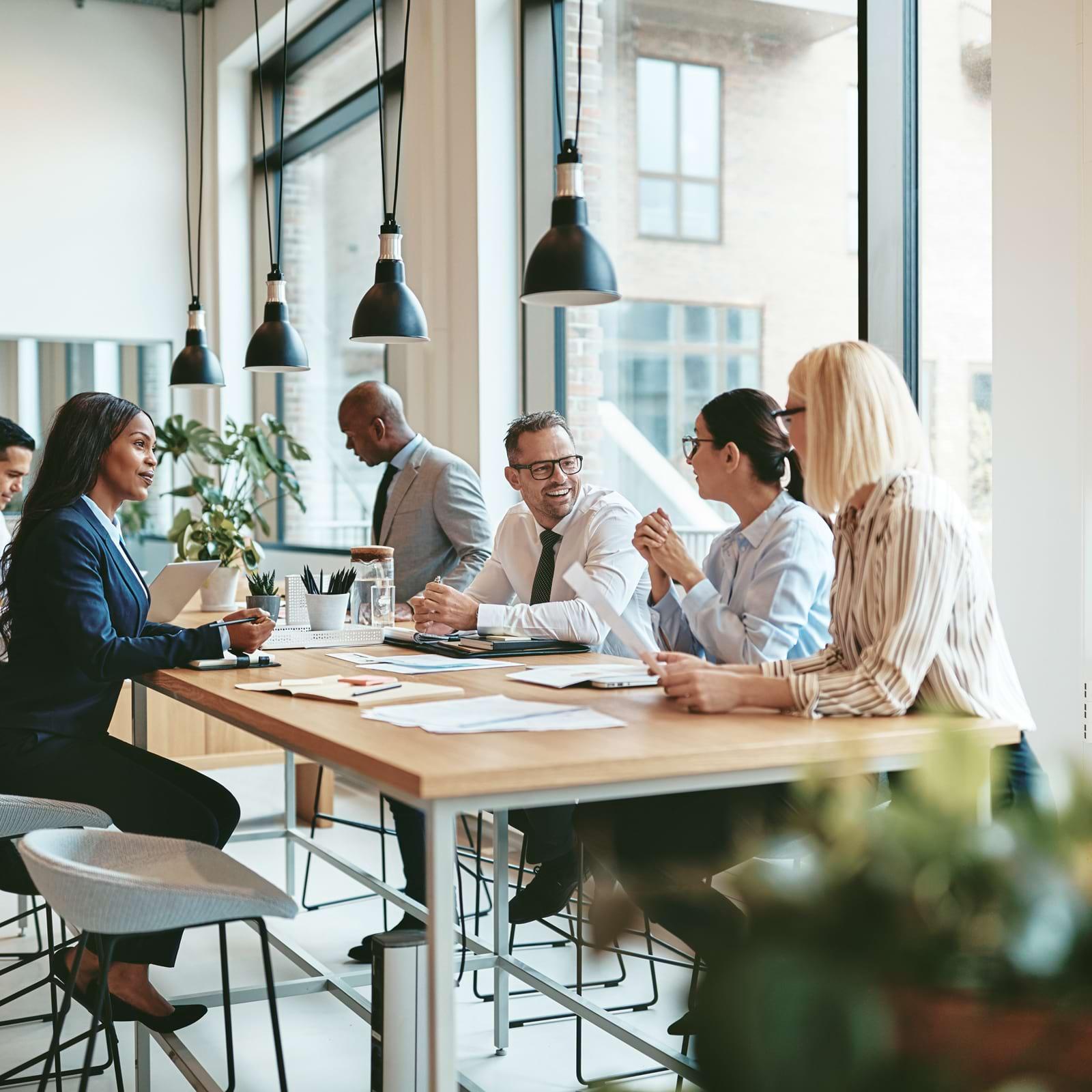 A diverse group of professionals engaged in a meeting around a wooden table in a modern office with large windows and hanging lights.