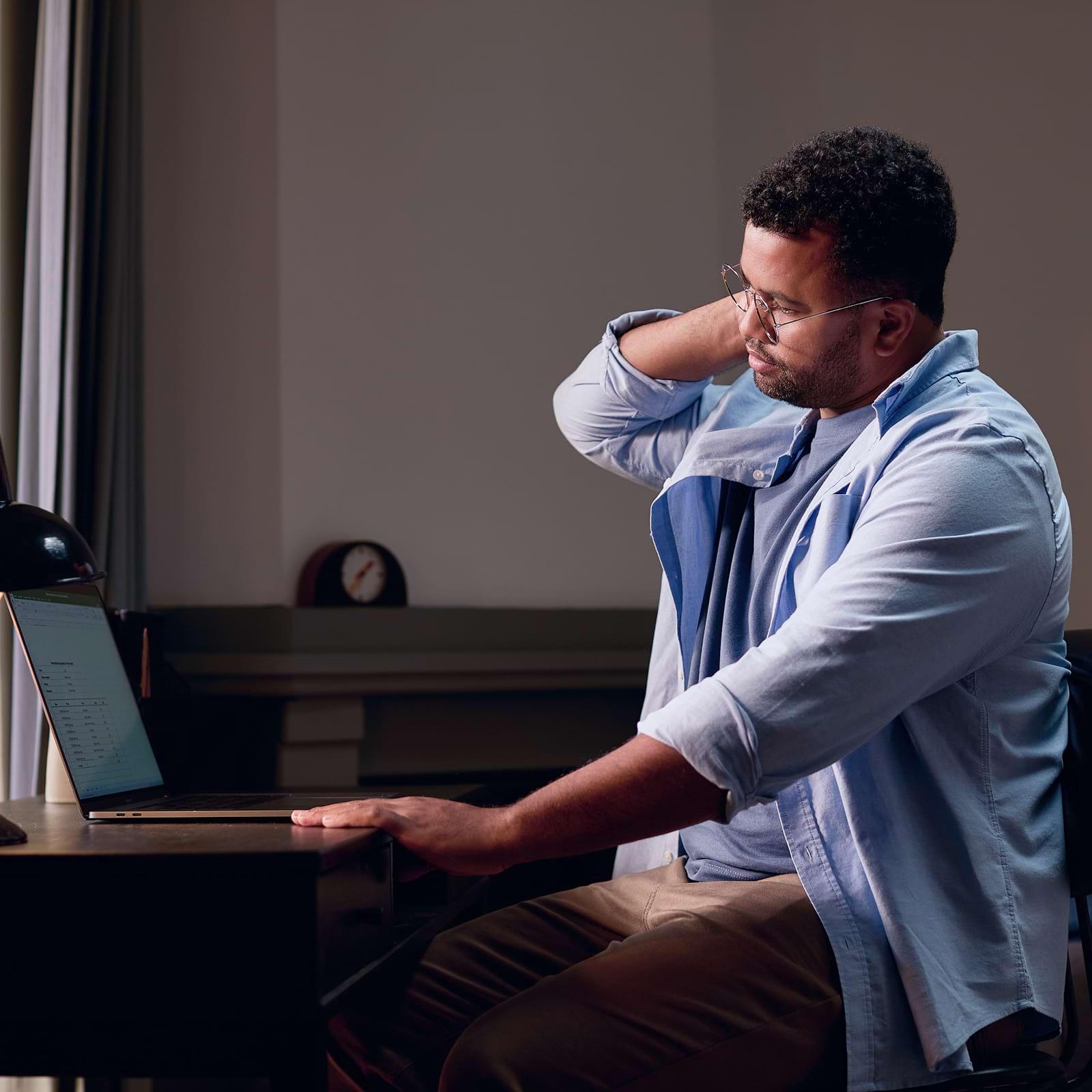 A man in a blue shirt sits at a desk, looking at a laptop and massaging his neck, appearing tired or tense.