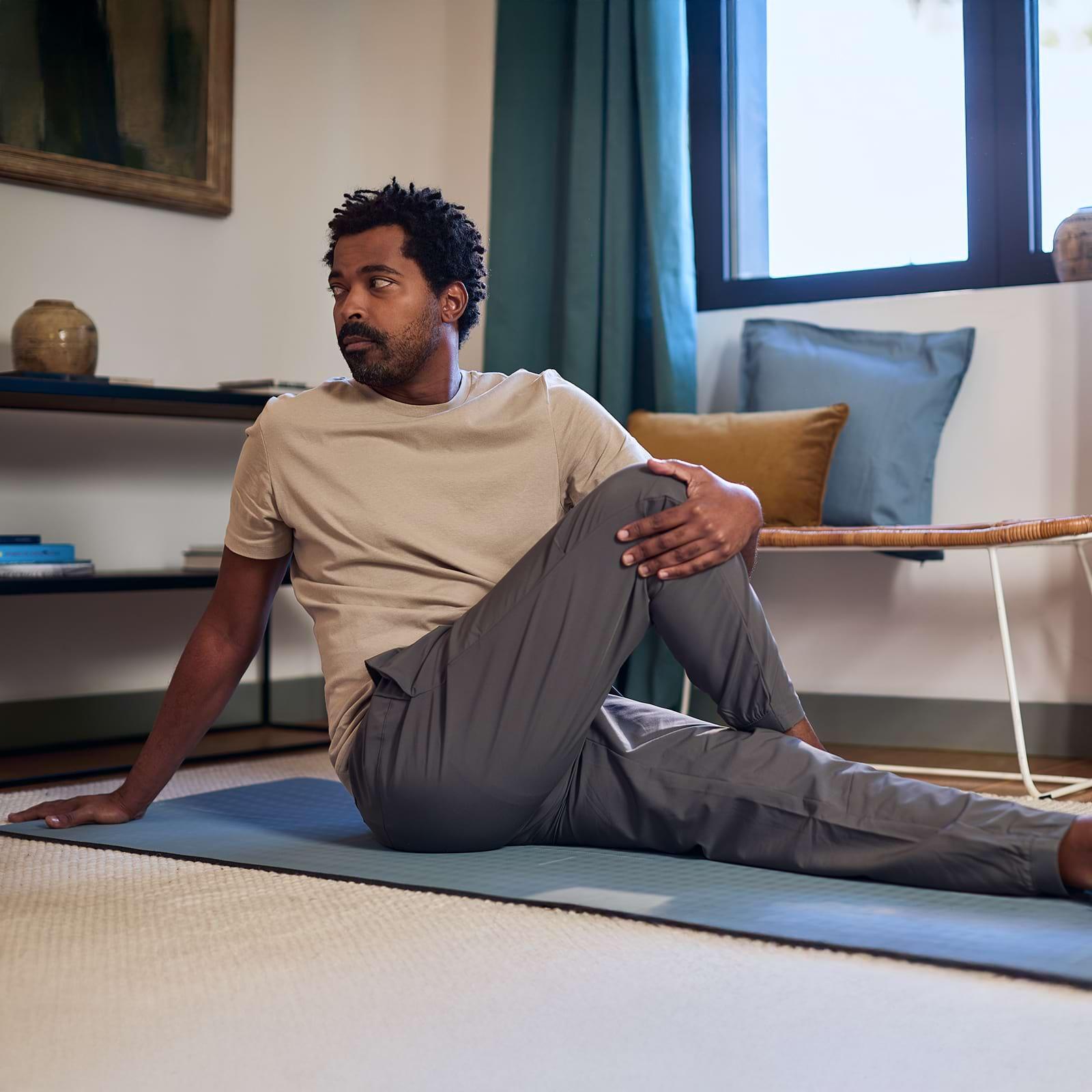Man in a beige t-shirt and gray pants doing a seated yoga twist on a mat in a cozy room with a window and cushions.