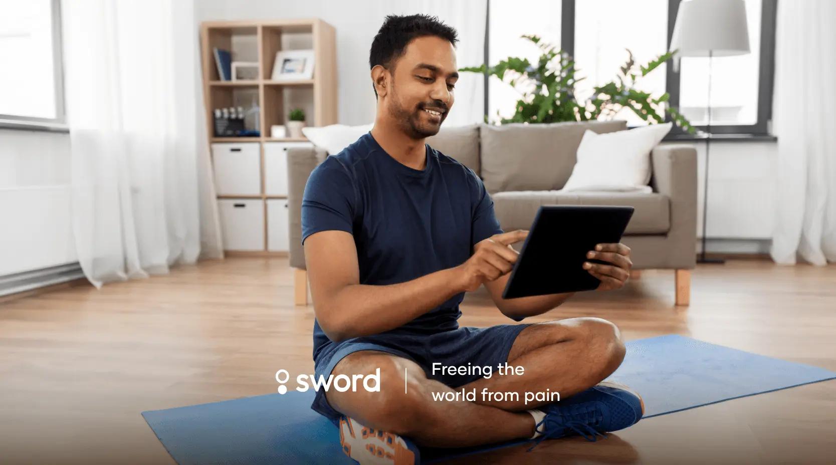 Man sitting cross-legged on a yoga mat, using a tablet, in a bright living room with modern decor.